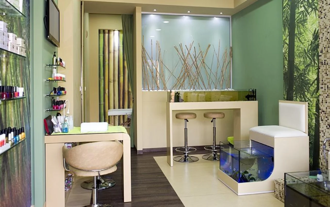 Un salón compacto con decoración mural de bambú y verde bosque, estanterías de pared con esmaltes de uñas, una mesa y silla de manicura color crema, dos taburetes de bar, y un acuario de cristal iluminado en azul para la pedicura fish spa.