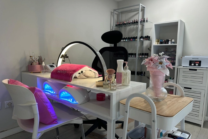 Puesto único de manicura con una mesa blanca, un aro de luz y dos lámparas LED con luz azul, una silla negra y estanterías cubiertas de esmaltes de colores, alegrado por un jarrón rosa con flores.