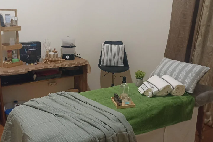 Cabina de masaje acogedora con una camilla cubierta por una toalla verde y toallas blancas enrolladas, una bandeja de aceites esenciales, herramientas de masaje de madera sobre una mesita rústica y un taburete junto a un rincón separado por una cortina.