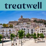 Vista soleada sobre Ibiza ciudad, con edificios encalados en blanco que suben hasta la catedral y las antiguas murallas de Dalt Vila en la colina. Un paseo marítimo arbolado atraviesa el primer plano bajo un cielo azul limpio.