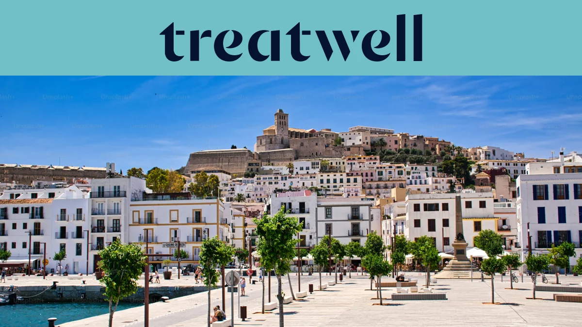 Vista soleada sobre Ibiza ciudad, con edificios encalados en blanco que suben hasta la catedral y las antiguas murallas de Dalt Vila en la colina. Un paseo marítimo arbolado atraviesa el primer plano bajo un cielo azul limpio.