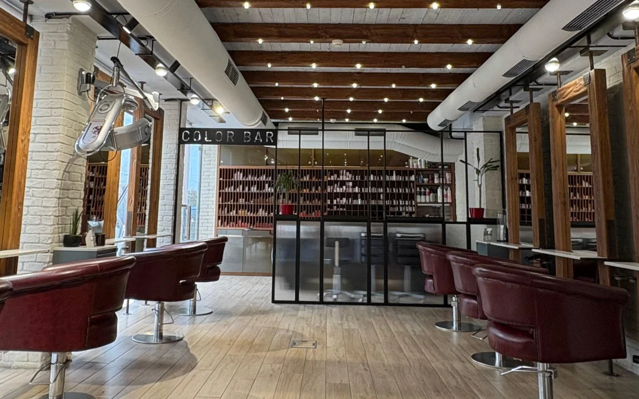 Un salón de peluquería con vigas de madera vistas y ladrillo encalado, sillones de estilismo de cuero burdeos, y una sala de color con paredes acristaladas forrada de estantes de productos detrás de un cartel 'Color Bar'.