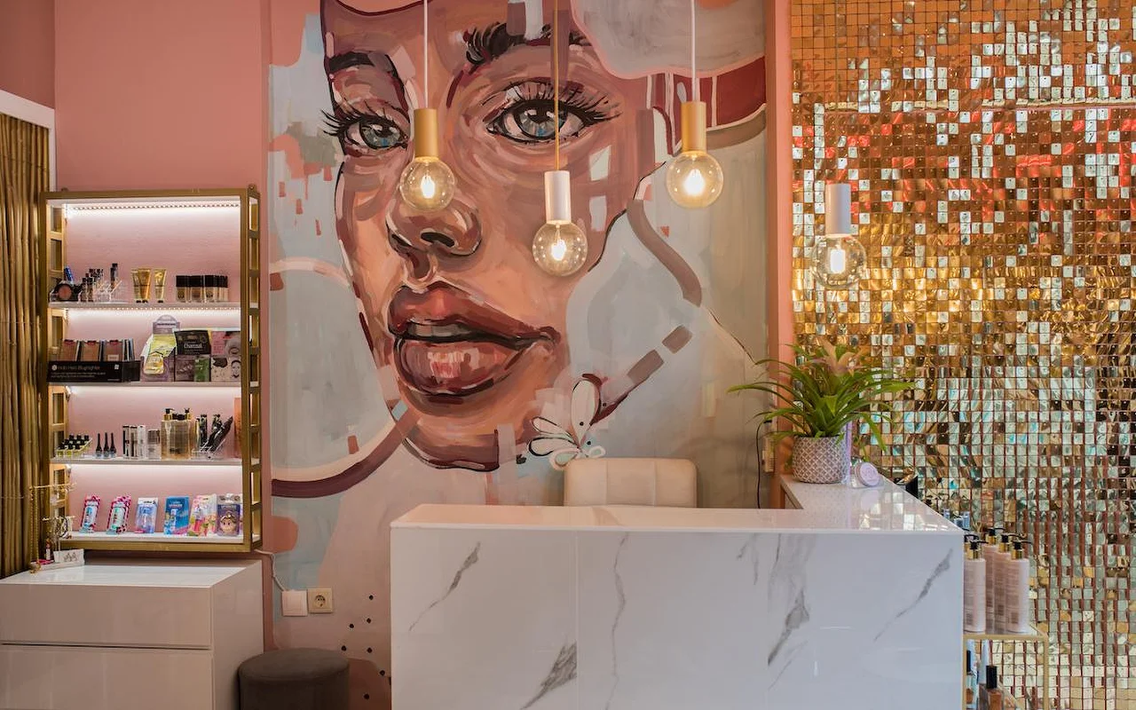 Un salón de paredes rosas con un gran mural pintado de un rostro de mujer, una pared de lentejuelas doradas brillantes, un mostrador de recepción de mármol blanco y lámparas colgantes con bombillas expuestas.
