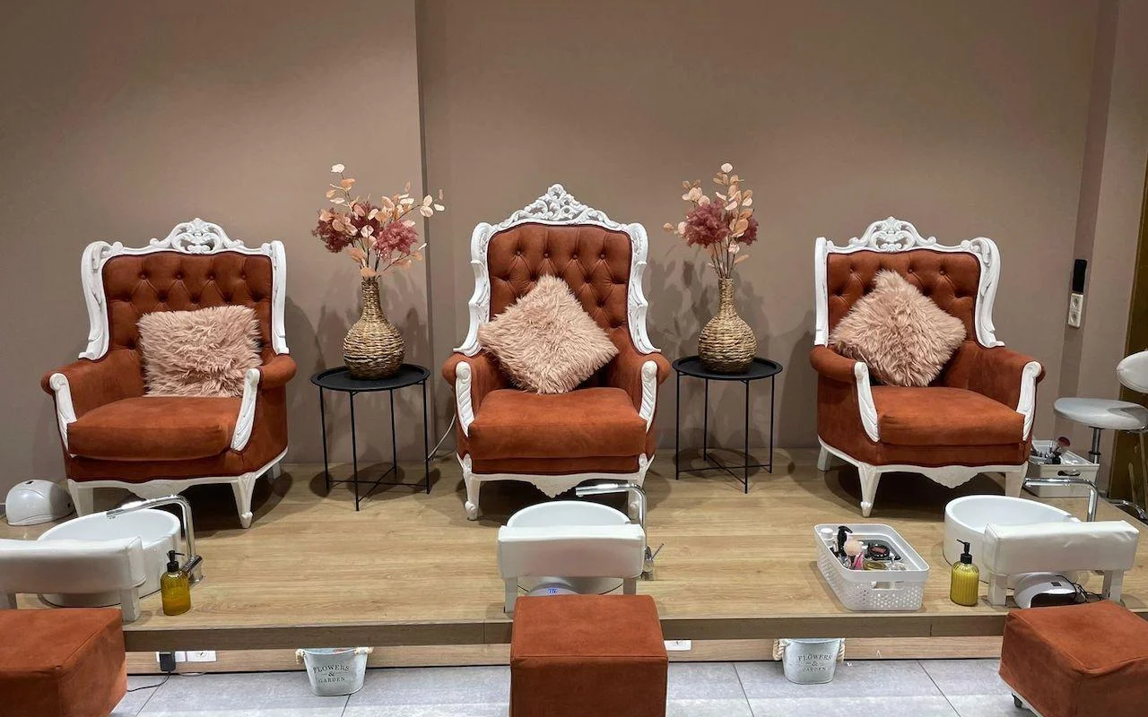 Tres sillones ornamentados con marcos blancos en terciopelo naranja quemado, elevados sobre una plataforma de madera encima de barreños de pedicura blancos, con flores secas y cojines rosa suave entre los asientos.