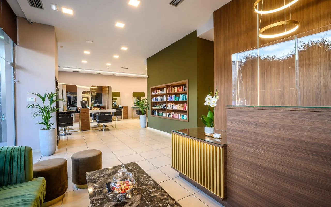 Una peluquería de alta gama con paneles de madera cálida, una pared decorativa verde oliva con estantes de productos, puestos de estilismo con grandes espejos, un mostrador de recepción con listones dorados y asientos de terciopelo verde bajo lámparas colgantes doradas circulares.