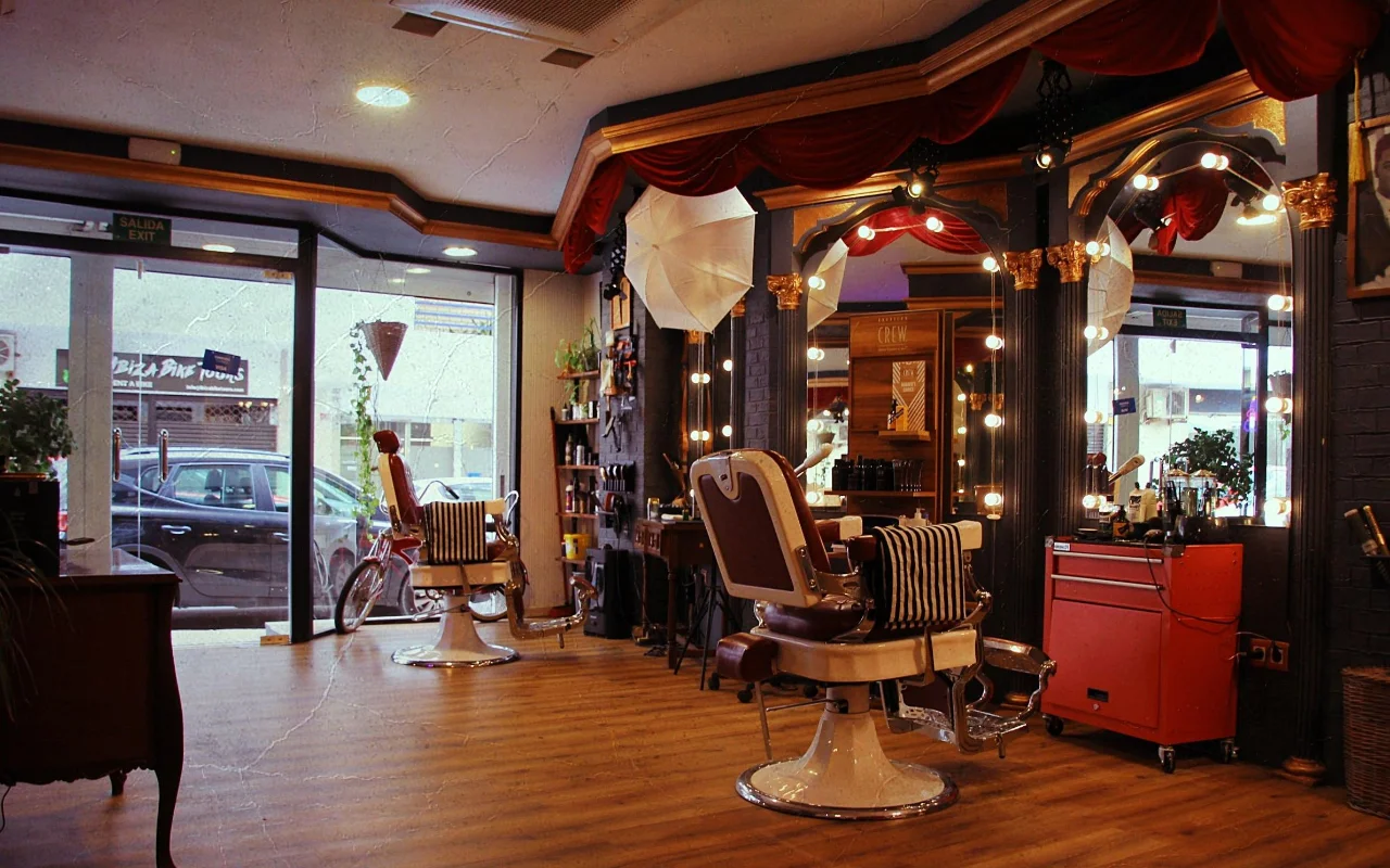 Una barbería de estilo vintage con clásicos sillones reclinables de cuero sobre bases cromadas y toallas de rayas. Espejos dorados enmarcados por bombillas redondas y cortinajes de terciopelo rojo recorren la pared, un carrito rojo de herramientas queda a un lado, y ventanas a la calle iluminan el suelo de madera.
