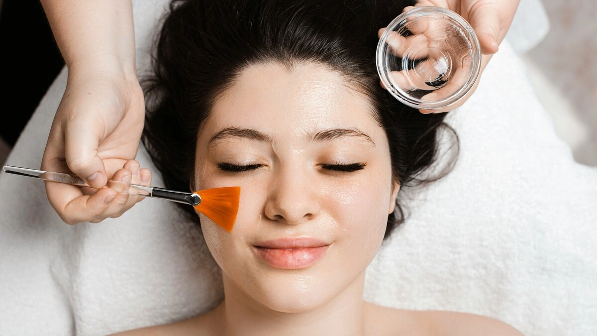 Vista cenital de una esteticista aplicando una solución de peeling químico en la cara de una clienta con una brocha ancha y plana