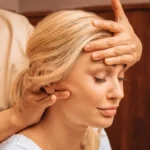 Primer plano de una mujer con los ojos cerrados mientras una profesional, durante un masaje indio de cabeza, le apoya una mano en la frente y con la otra le masajea el cuello