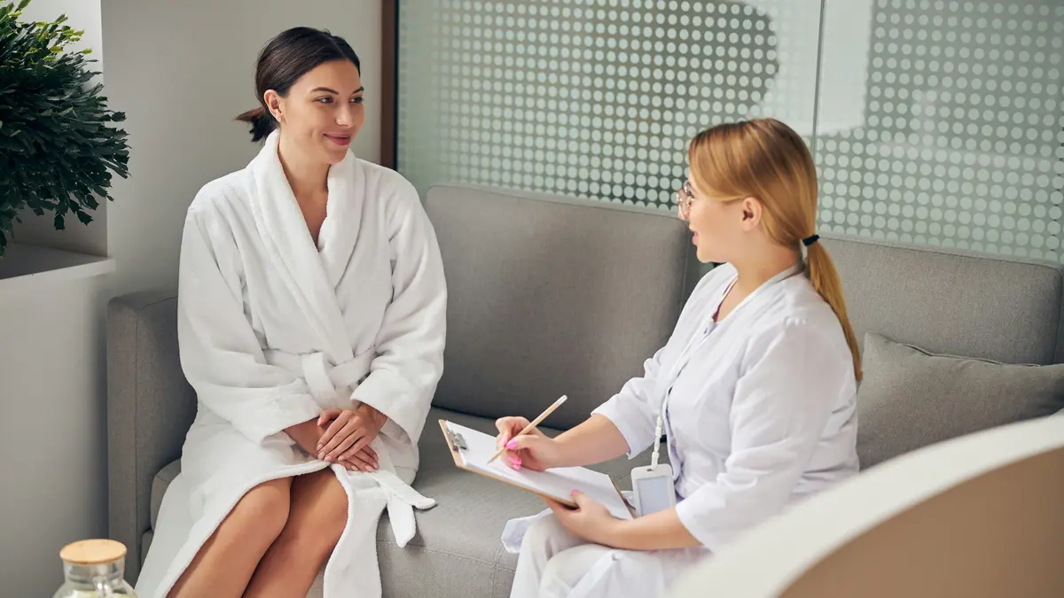 Una clienta con albornoz blanco y una profesional con un portapapeles, sentadas una al lado de la otra en un sofá de un centro de estética, mientras la profesional le hace las preguntas previas al tratamiento y anota las respuestas
