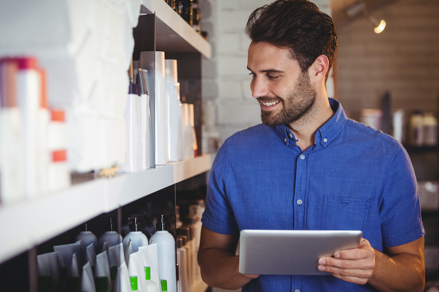 Un dueño de salón sonriente con camisa azul mira una tableta junto a estanterías de productos de peluquería y belleza.