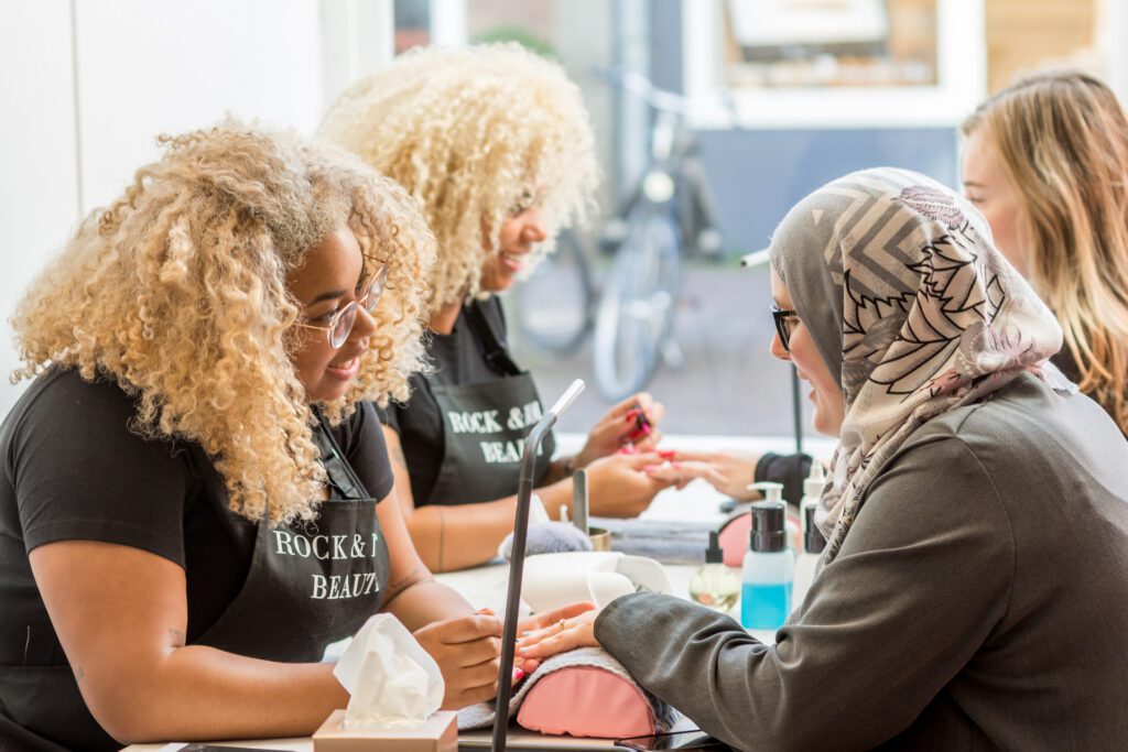 Dos manicuristas de pelo rubio rizado con delantales negros donde se lee "Rock & Roll Beauty" hacen la manicura a unas clientes en una mesa larga. Una cliente lleva un hiyab gris.