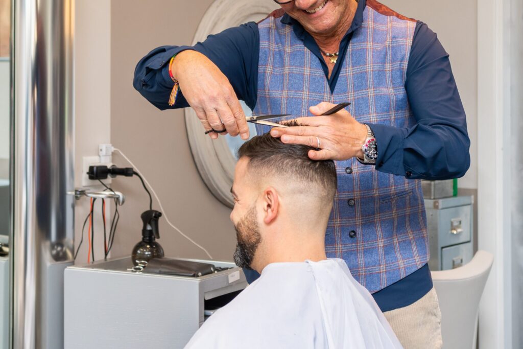 Un peluquero con un chaleco de cuadros azules corta el pelo a un cliente utilizando tijeras y un peine en un salón. El cliente tiene el pelo corto, barba y lleva una capa de corte blanca.
