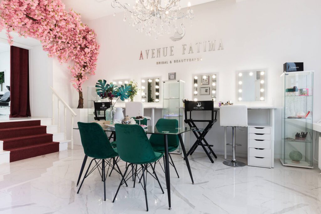 Un luminoso y elegante salón de belleza y novias llamado "Avenue Fatima". El espacio cuenta con varias estaciones de maquillaje con espejos iluminados, sillas de terciopelo verde esmeralda alrededor de una mesa de vidrio y un cerezo rosa decorativo.