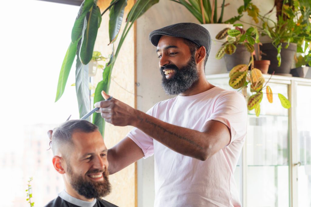 Un barbero sonriente con barba oscura y una gorra plana gris utiliza tijeras y un peine en el cabello de un cliente sonriente en un salón muy iluminado y lleno de plantas verdes.