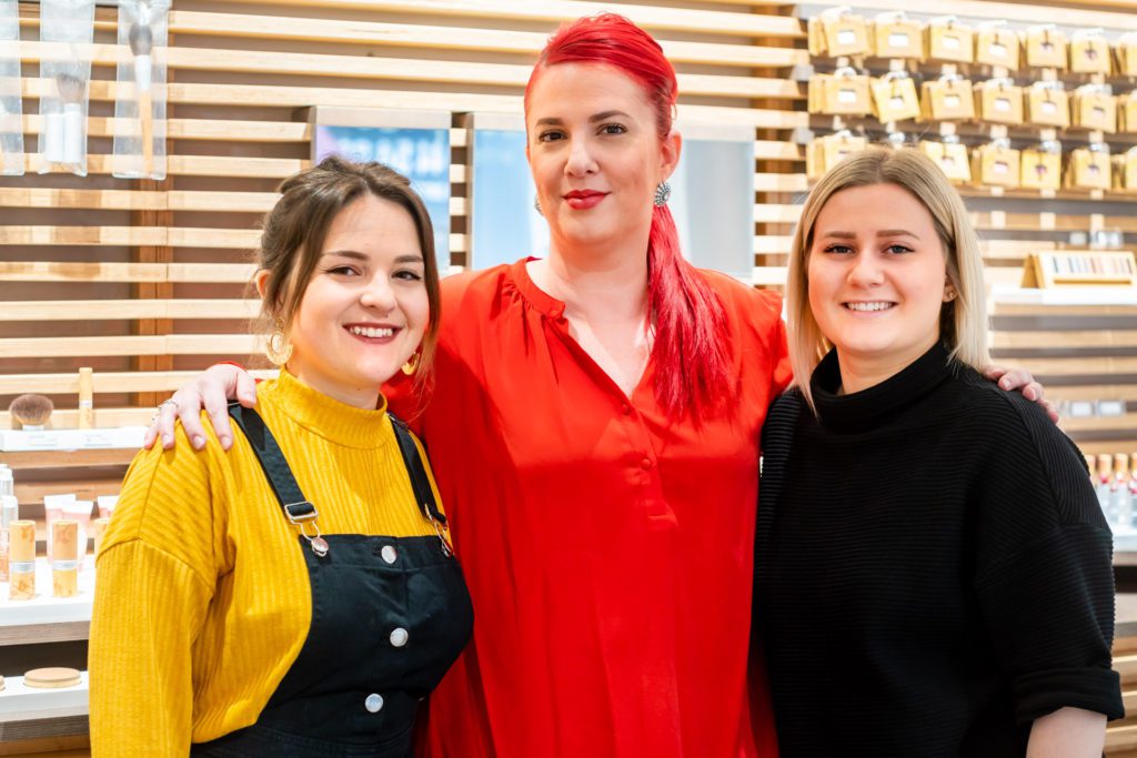 Tres profesionales de salón sonrientes posan juntas abrazadas frente a una pared de listones de madera que exhibe productos cosméticos. La mujer del centro tiene el cabello rojo brillante y una blusa roja.