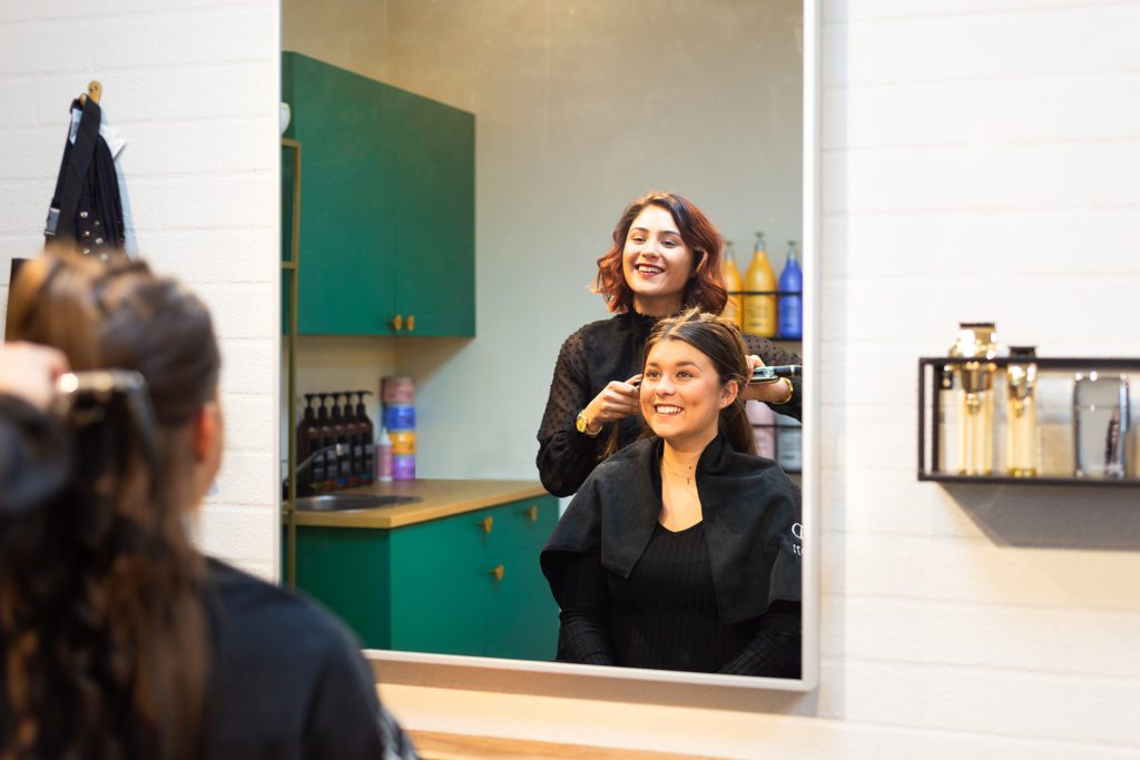 Una peluquera sonriente con cabello castaño rojizo moldea el pelo de una cliente sonriente con una plancha, vista a través del reflejo de un gran espejo rectangular en un salón moderno con muebles verde esmeralda.