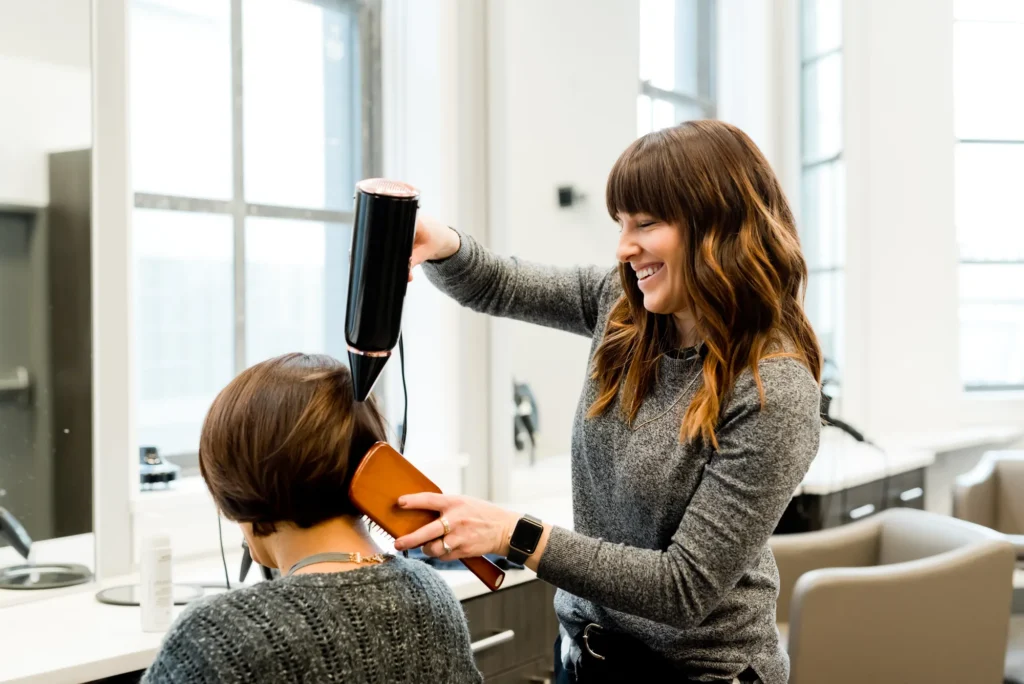 Una peluquera sonriente seca y cepilla el pelo de un cliente en un salón luminoso y moderno.